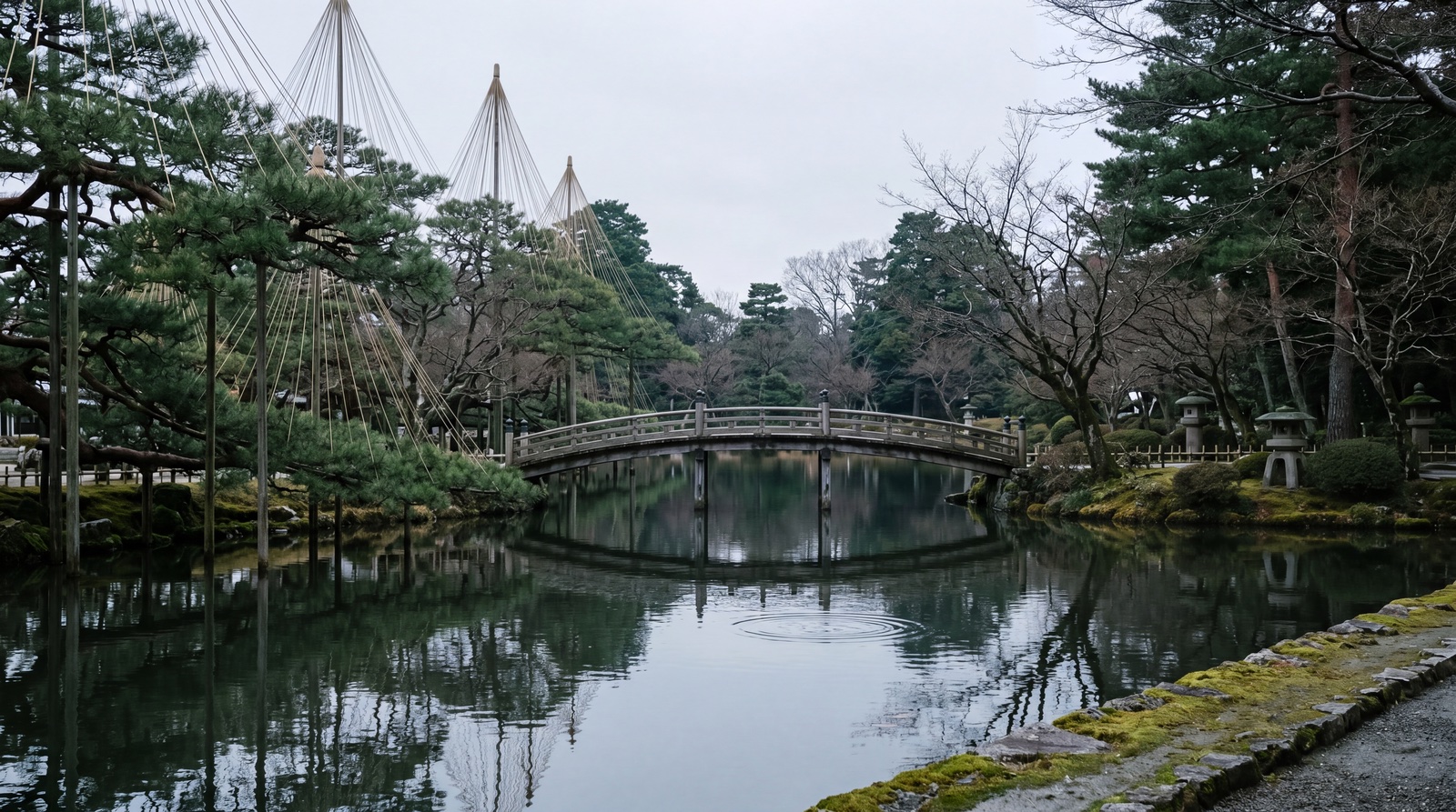 Kenroku-en garden in early March. A wooden footbridge over still dark water, pine trees with their winter yukitsuri snow ropes — Kanazawa in its quiet season.