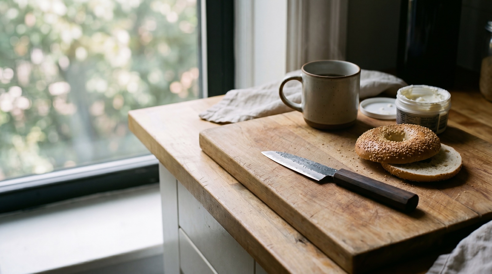 A quiet Brooklyn kitchen counter on a Saturday morning, weeks after the trip. A small handmade kogatana rests on a wooden cutting board next to a sliced sesame bagel and a steaming coffee mug.